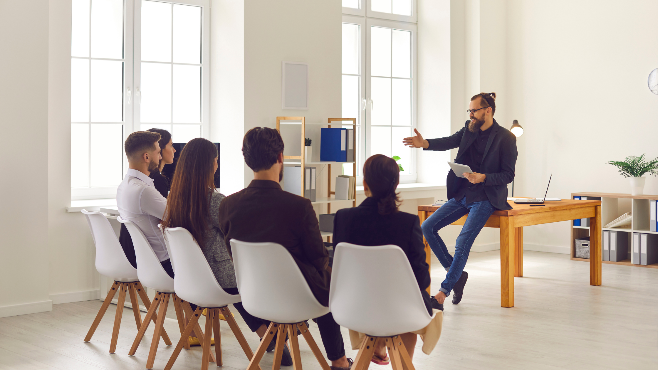 Five business professionals sit on chairs in a line listening to a male who is sitting on a table holding a tablet, potentially discussing leadership development strategies.